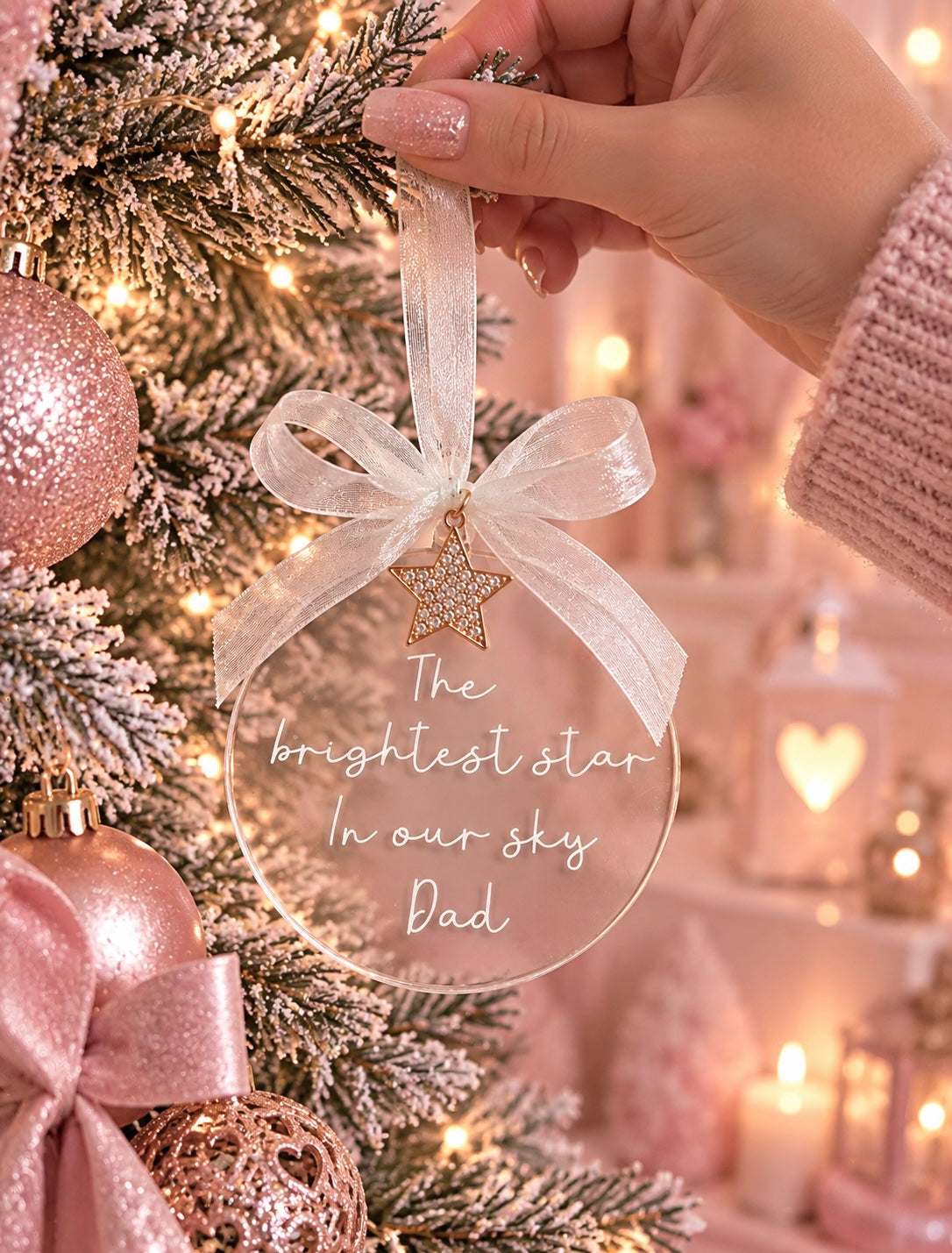 A hand holds a clear round Christmas ornament with a white ribbon bow and a gold star, printed with the message 'The brightest star In our sky Dad'. The ornament is displayed on a decorated Christmas tree with pink ornaments and warm fairy lights.