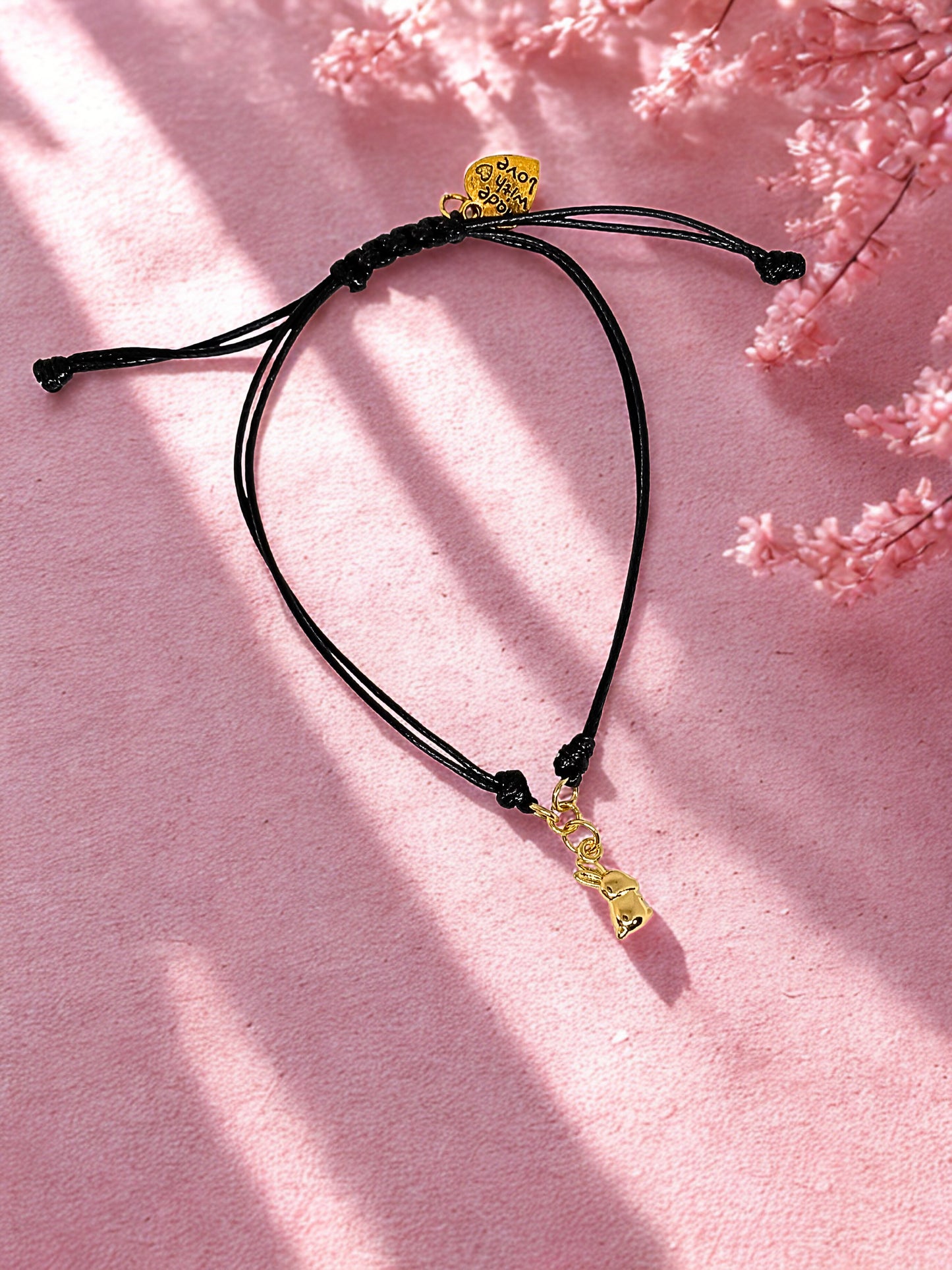 Black cord bracelet with gold charm on a pink background
