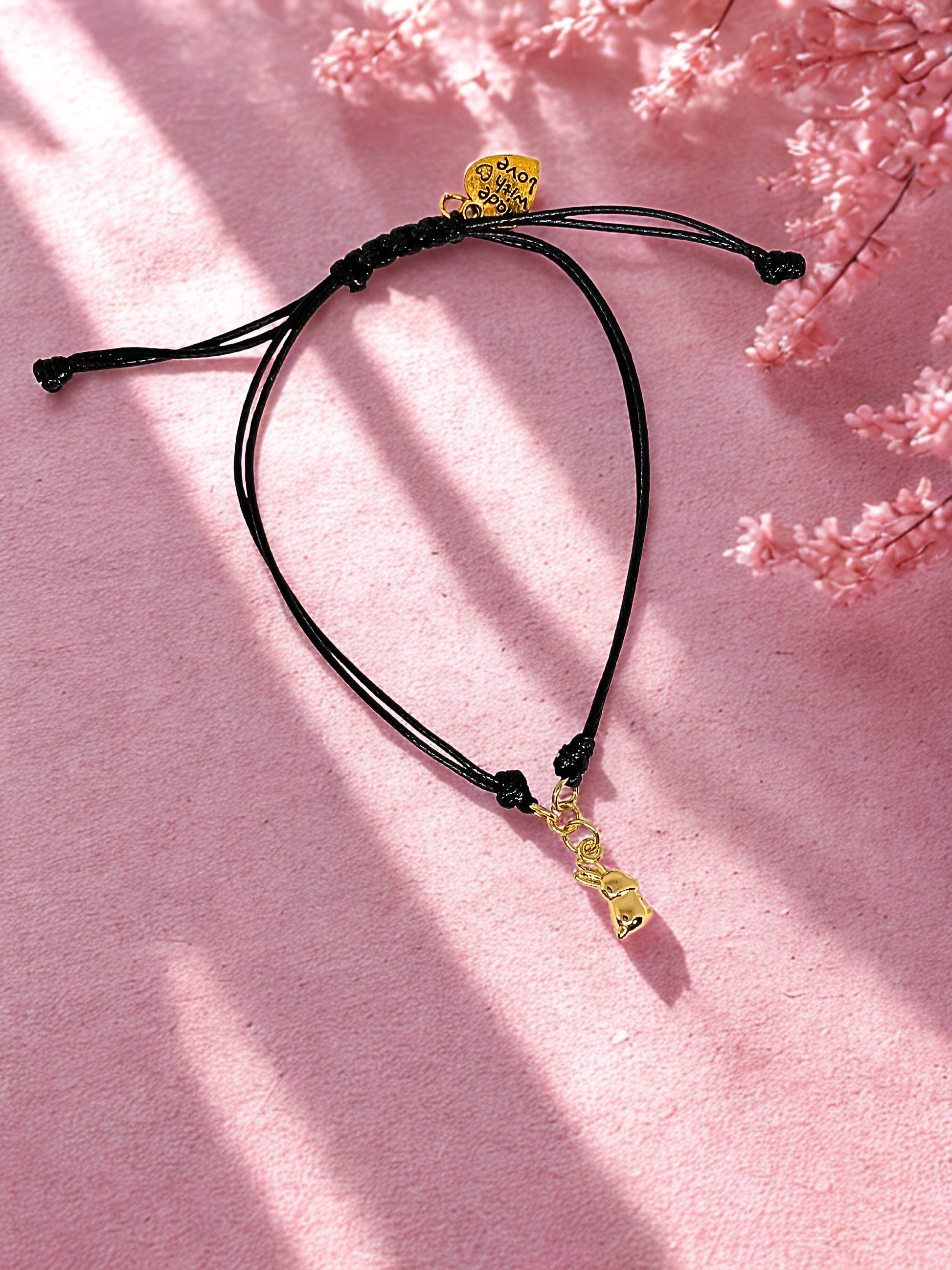 Black cord bracelet with gold charm on a pink background