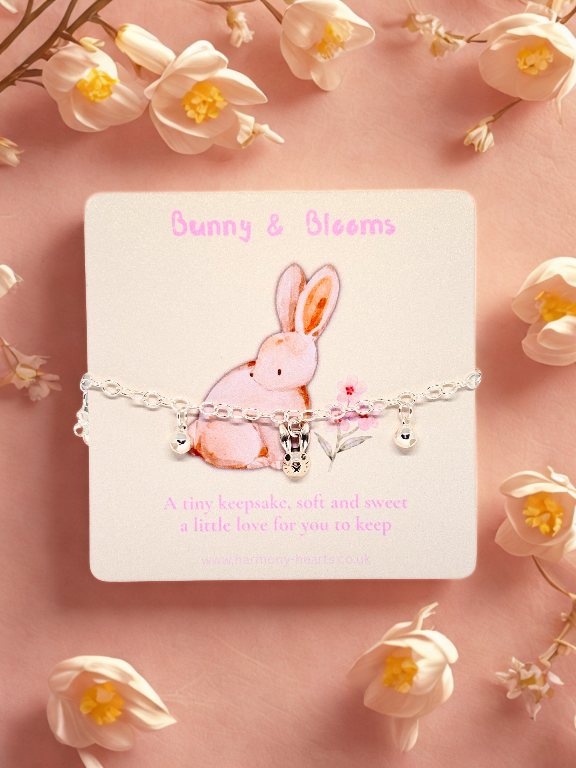 Bracelet with a bunny charm on a card labeled 'Bunny & Blooms' surrounded by white flowers on a pink background.