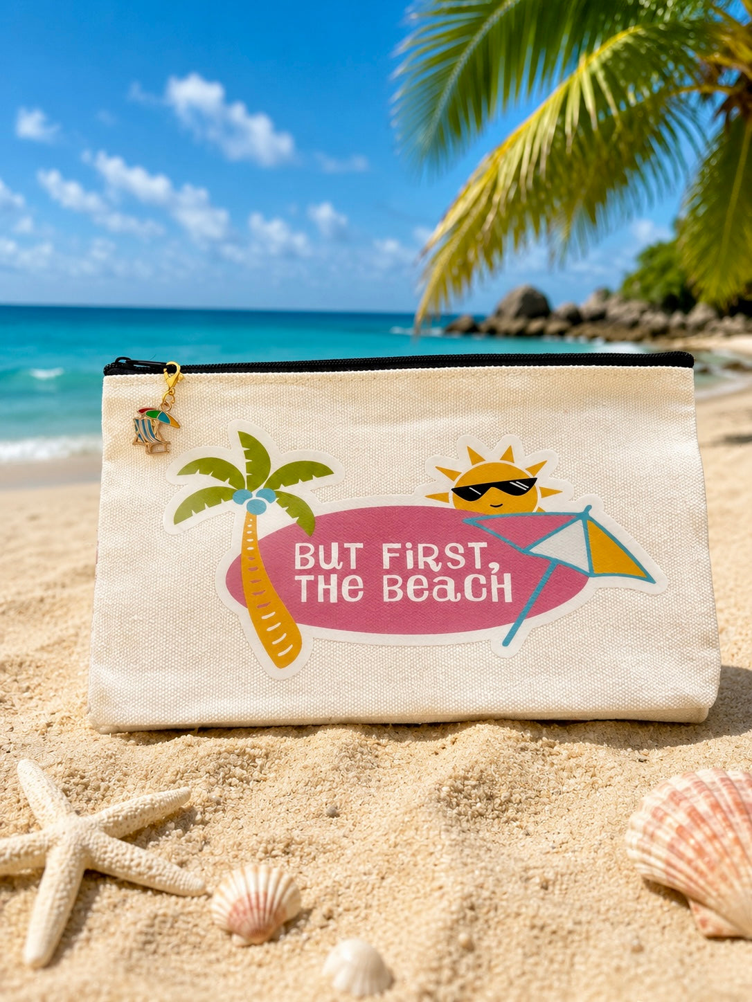 Beach pouch with 'But First, The Beach' text and palm tree design on a sandy background with ocean and palm trees.