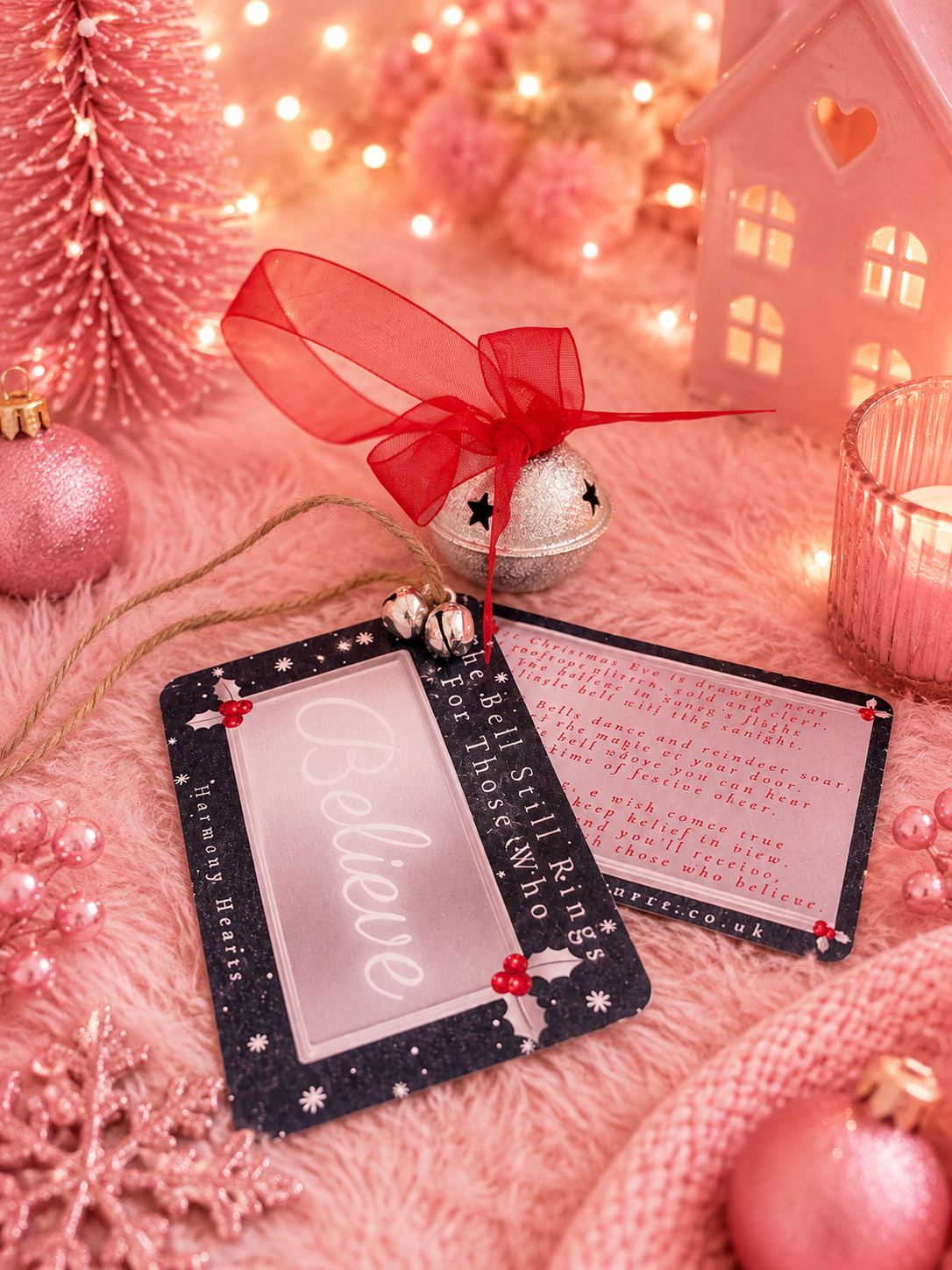 A festive Christmas believe bell set styled on a pink faux fur surface with holiday decorations. One card is closed, showing a glittery dark navy border and the word 'Believe' in white script on a pink background.