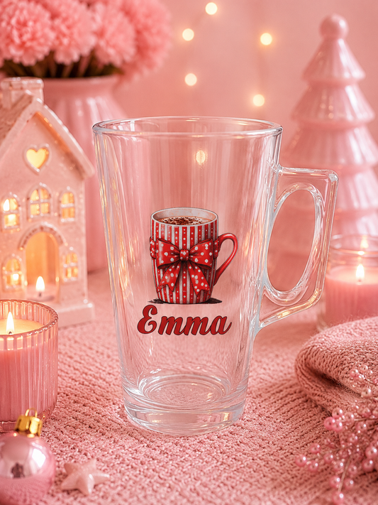 A clear glass mug with a handle, decorated with an illustration of a red mug with a white polka-dot bow and the name 'Emma' in red script, set against a cozy pink holiday background with candles and lights.