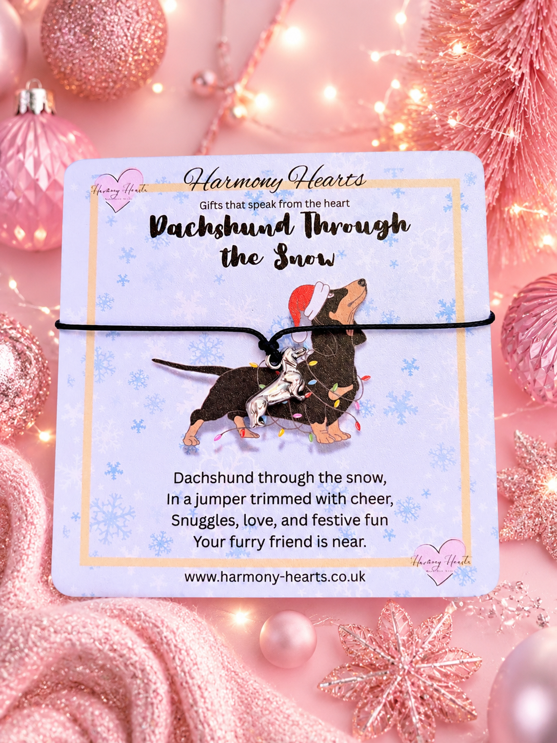 A charm bracelet on a card featuring a dachshund and a small dog wearing a Santa hat. The card reads 'Harmony Hearts' and 'Dachshund through the Snow' with a website at the bottom.