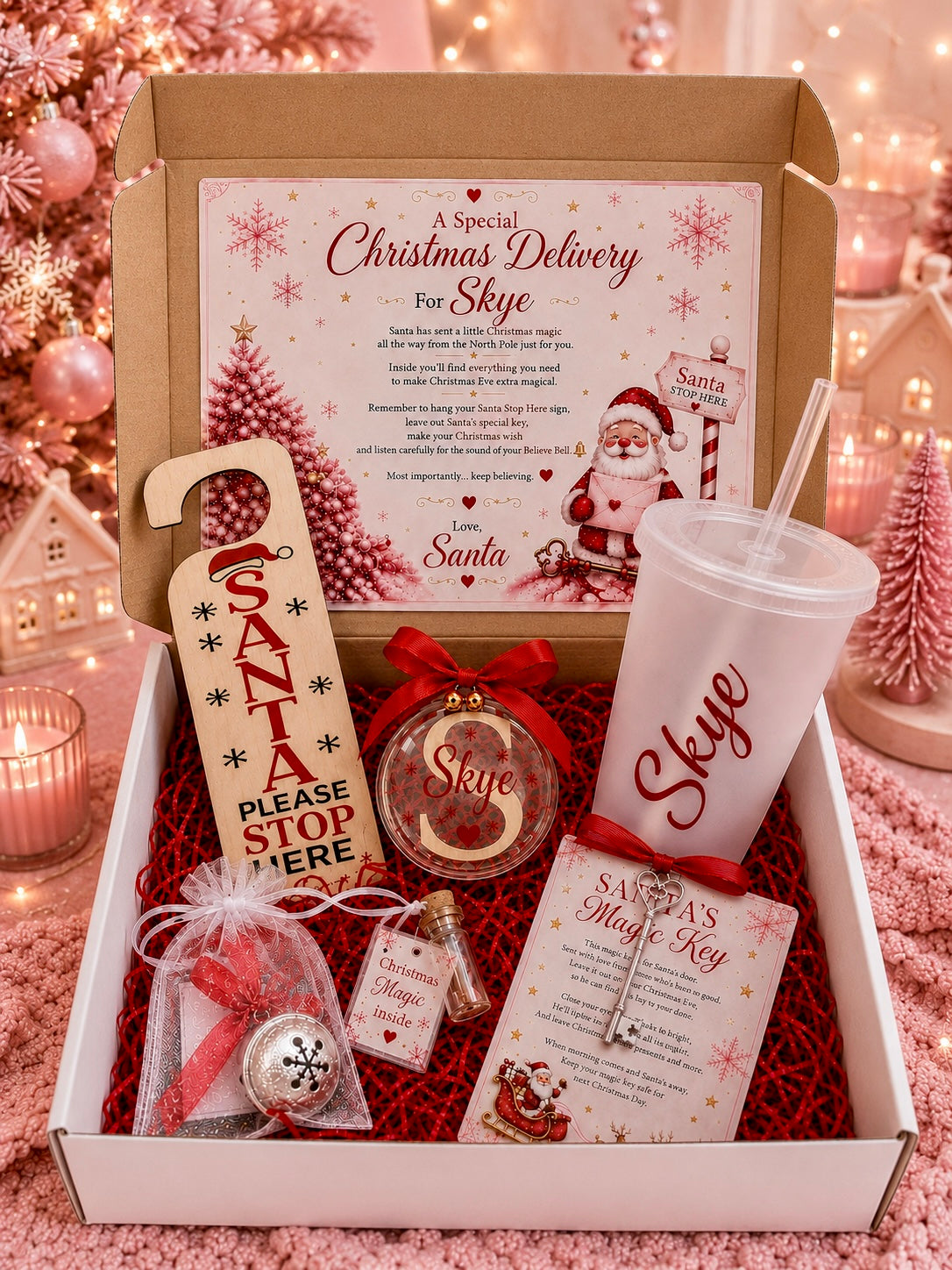 A curated Christmas gift box displayed open on a pink textured surface, filled with holiday-themed items including a printed Santa message card, a clear tumbler with red 'Skype' lettering, a red ribbon ornament, a 'Santa Please Stop Here' door hanger, a small gift bag, and a key. The box is surrounded by warm holiday decor, including a blurred Christmas tree and lights.