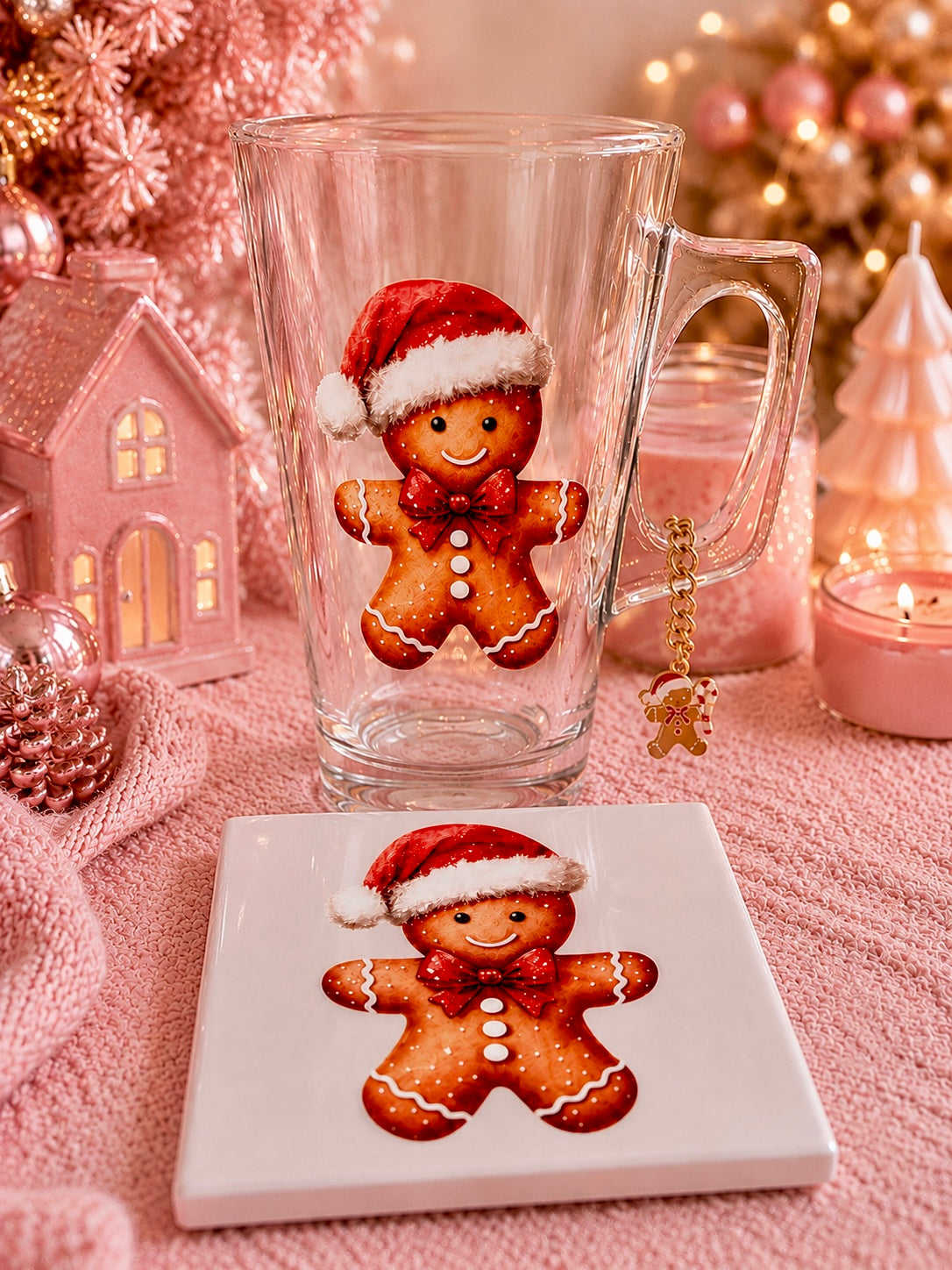 A clear glass mug with a gingerbread man illustration wearing a red Santa hat, shown beside a square white coaster with a matching gingerbread man design. The scene is styled in a cozy holiday setting with pink candles, ornaments, and a blurred Christmas tree in the background.