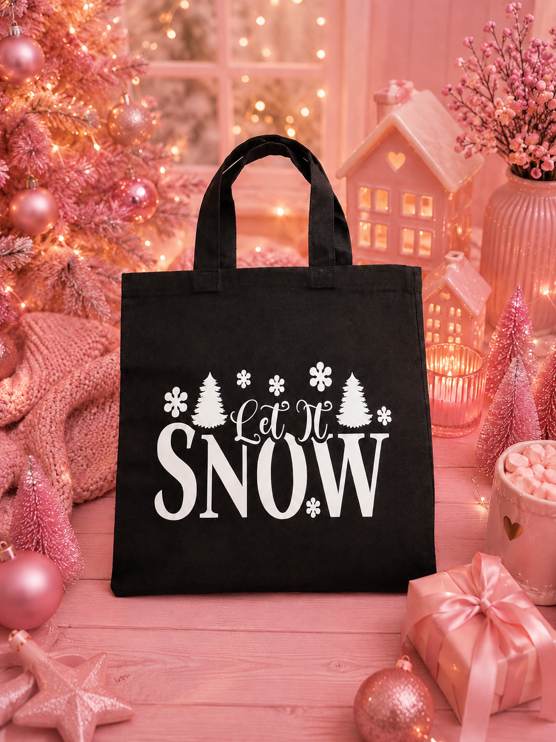 A black canvas tote bag with white 'Let It SNOW' text and snowflake/tree graphics, displayed on a pink holiday-themed surface with lights, ornaments, and gift boxes.