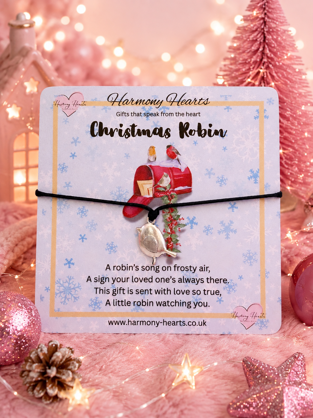 A Christmas Robin themed charm on a black cord, presented on a branded Harmony Hearts card. The card is styled in a festive, warm pink holiday setting with miniature Christmas trees, a pinecone, and warm bokeh lights.