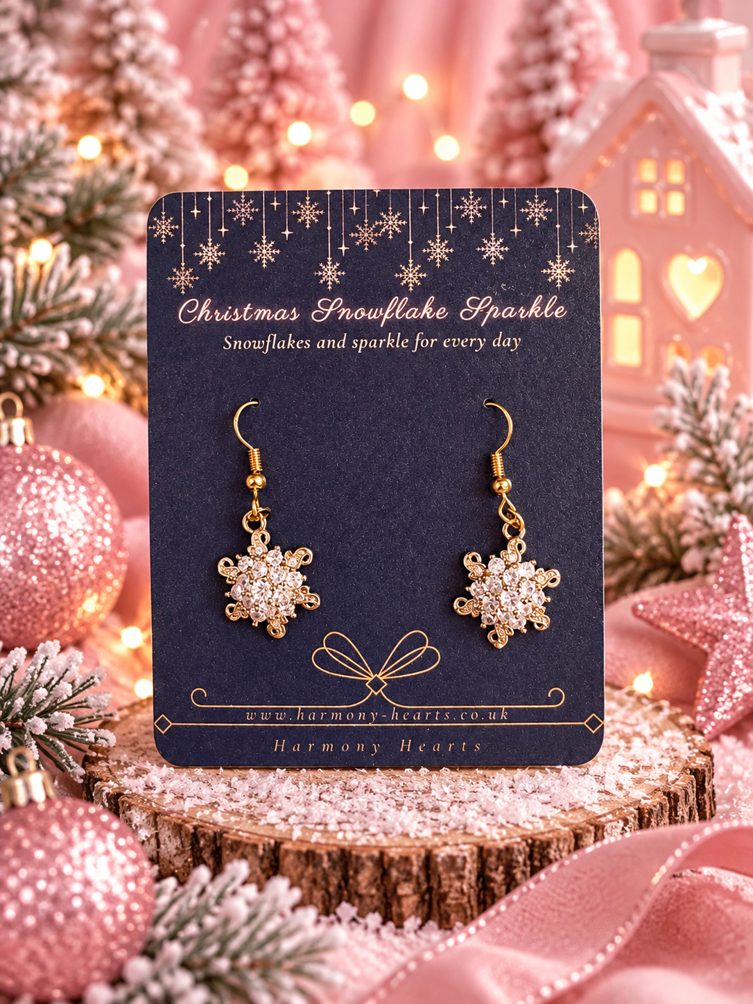 A pair of gold snowflake drop earrings with sparkling clear stones, displayed on a dark navy blue card. The card is set against a festive Christmas background with pink ornaments, lights, and a wooden slice.