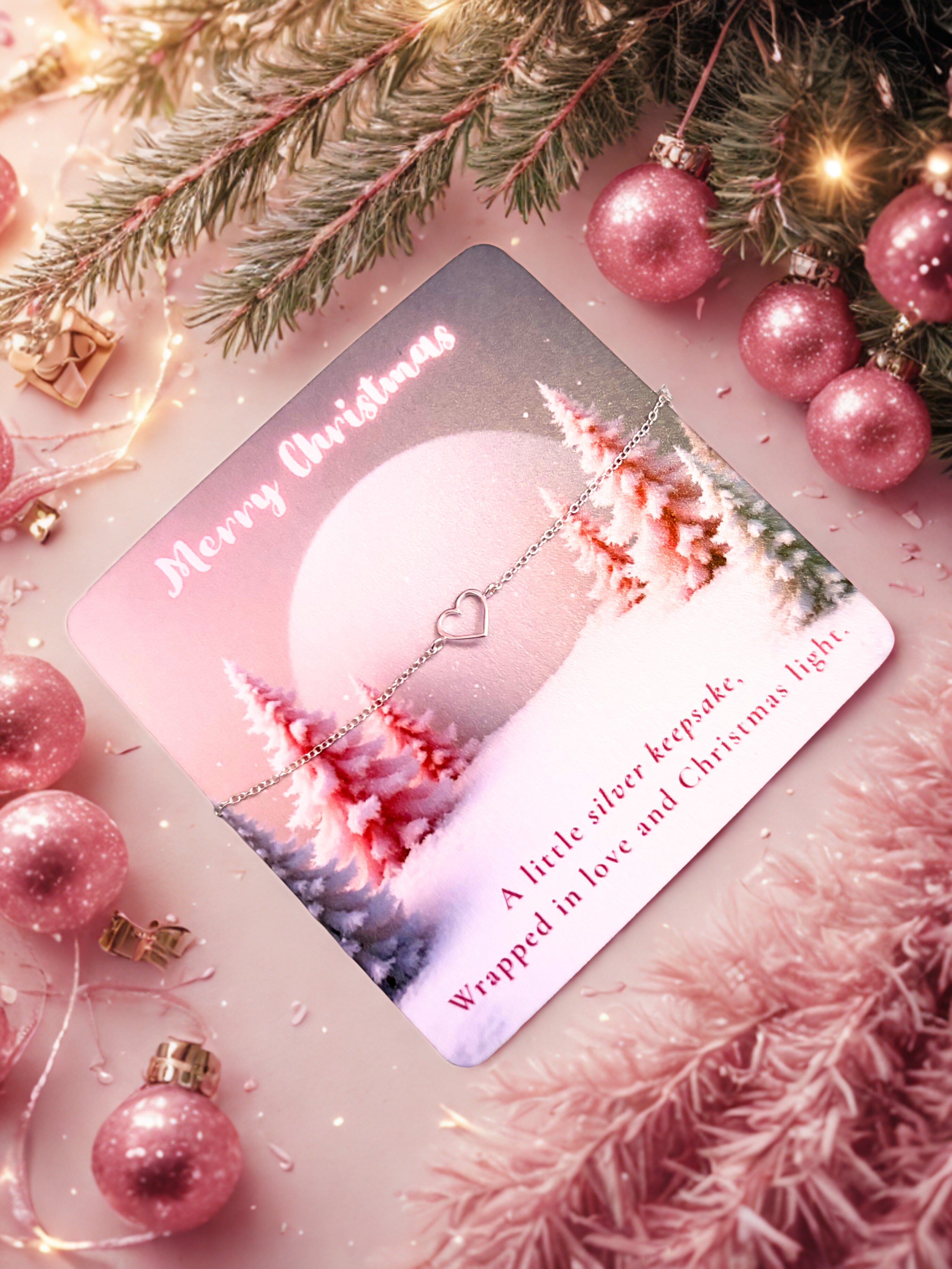 Christmas card with a silver heart bracelet, surrounded by pink ornaments and tree branches.