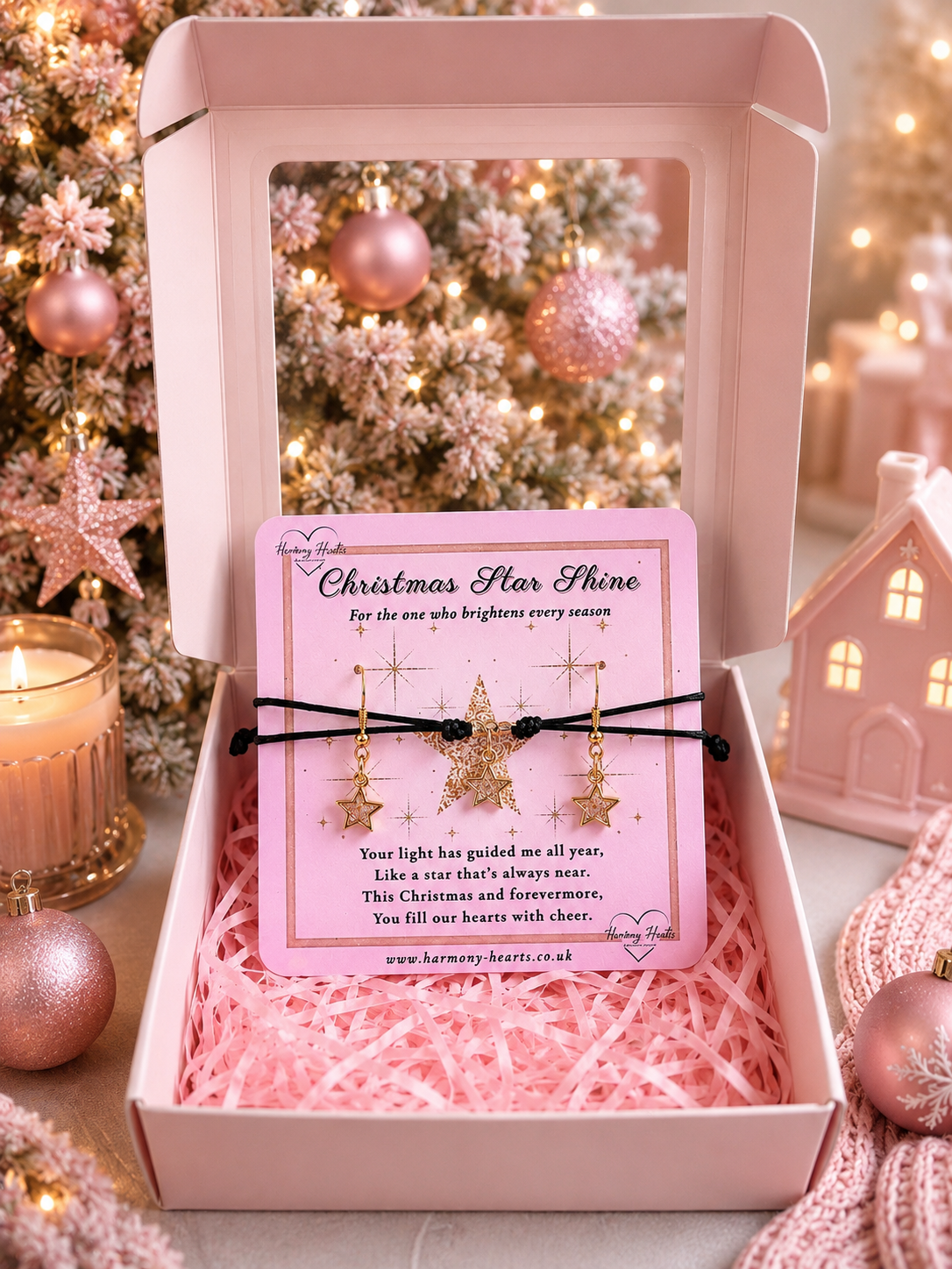 A pink gift box with a clear window displays a pink card reading 'Christmas Star Shine' with a black cord bracelet and a gold star charm and matching earrings. The box is filled with pink shredded paper, with a lit candle, pink ornaments, and a small house decoration in the background.