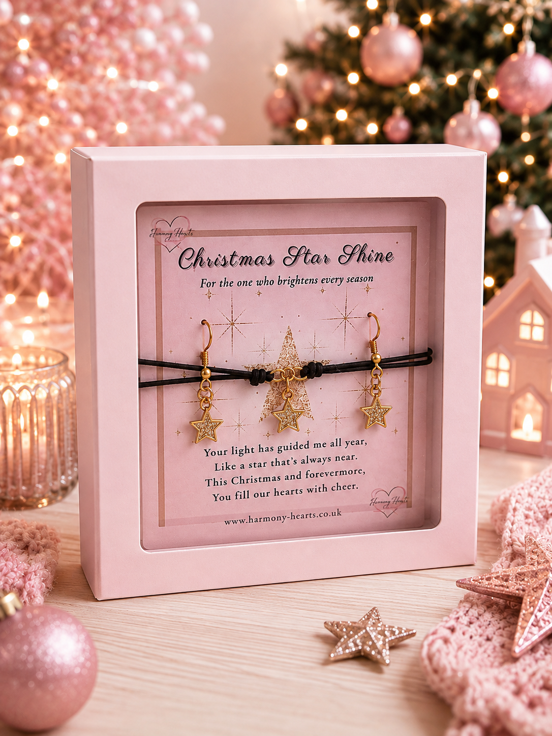 A pink gift box containing a Christmas-themed black cord bracelet with a star charm and with two gold star earrings, displayed on a wooden surface with festive pink ornaments and warm lights in the background.