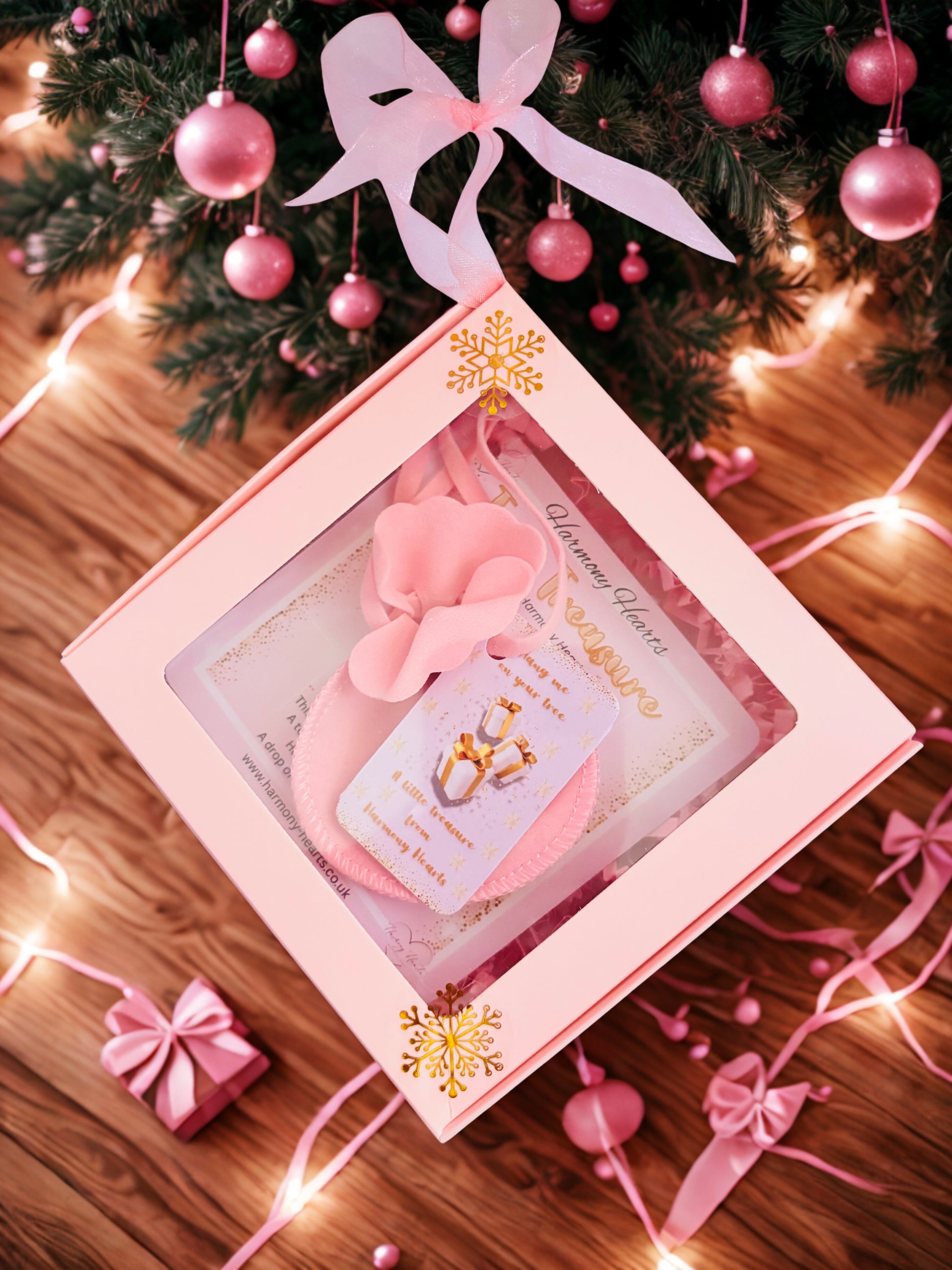 Pink gift box with decorative elements on a wooden surface with Christmas tree and lights.