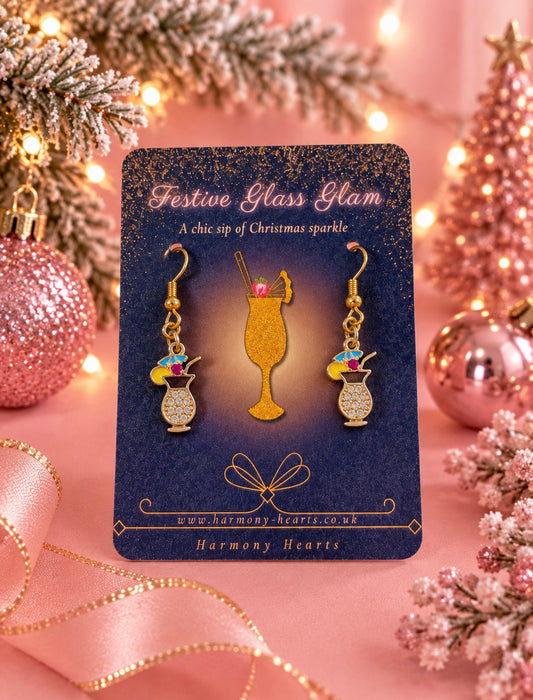 A pair of gold-tone cocktail-themed dangle earrings displayed on a dark blue card labeled 'Festive Glass Glam' by Harmony Hearts. The earrings hang from a gold drink glass charm and feature small cocktail vases with colorful umbrellas and straws.