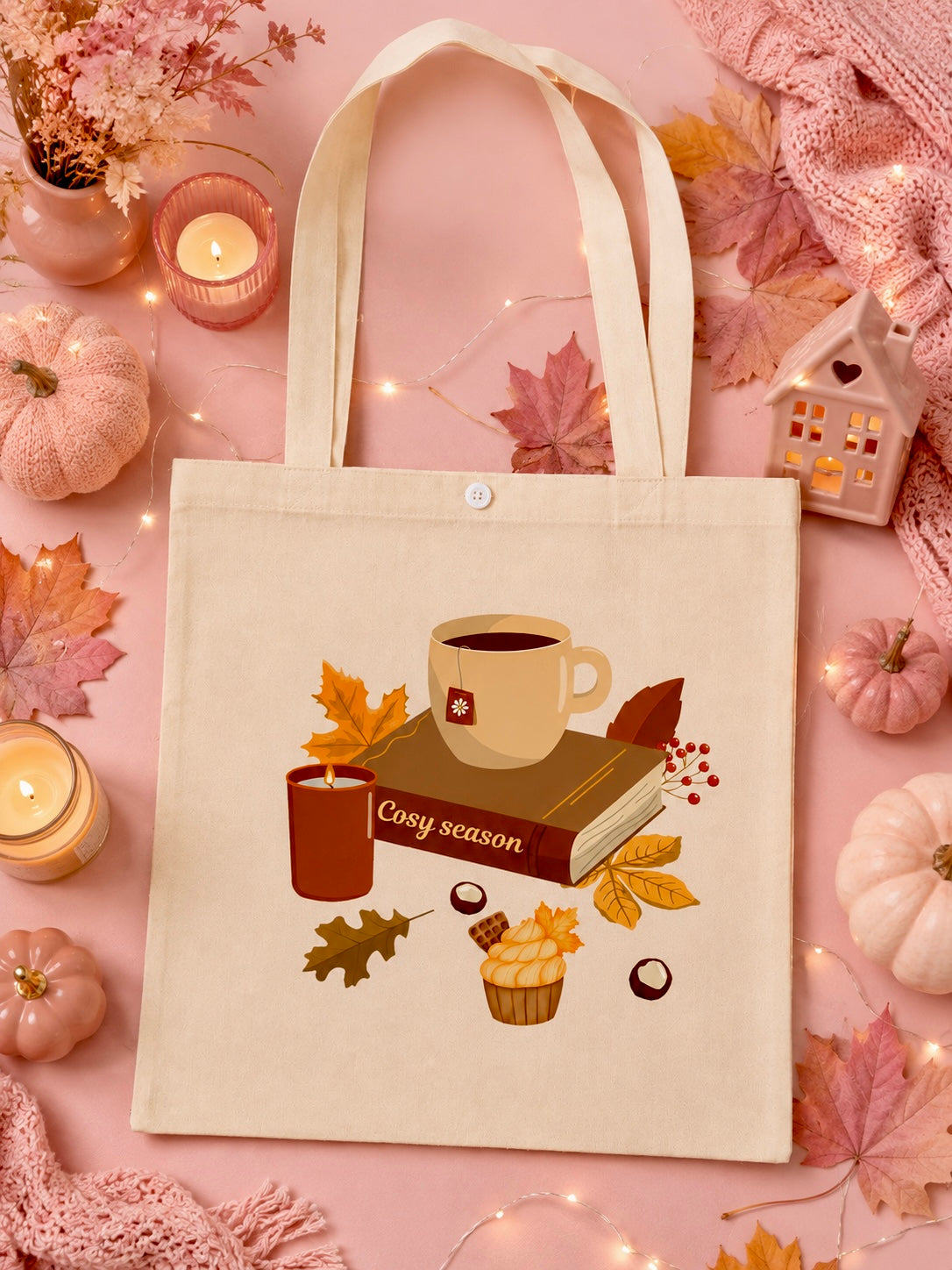 A cream canvas tote bag with a cozy autumn-themed illustration of a mug, book, candle, and cupcake with the text ' cosy season.' It is styled in a flat lay on a pink background with pumpkins, leaves, candles, and soft home decor.