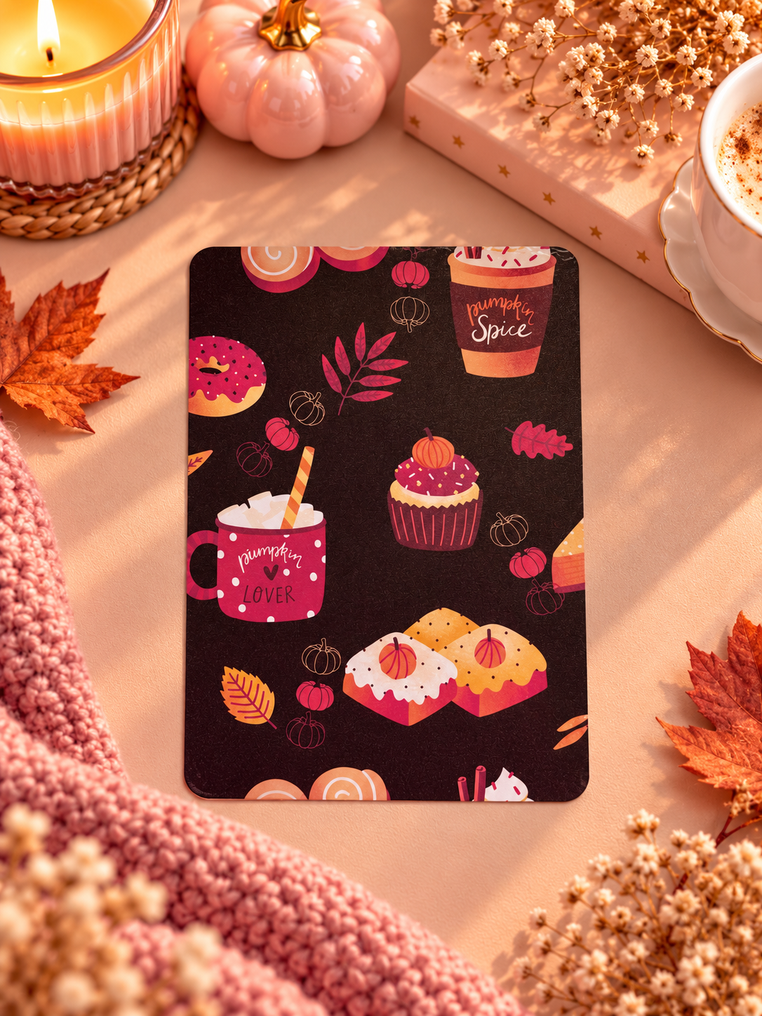 A rectangular black kindle insert with a fall-themed pattern of cupcakes, donuts, hot cocoa cups, and pumpkins, styled on a beige surface with autumn leaves, a candle, a wrapped gift, and a pink knit blanket.