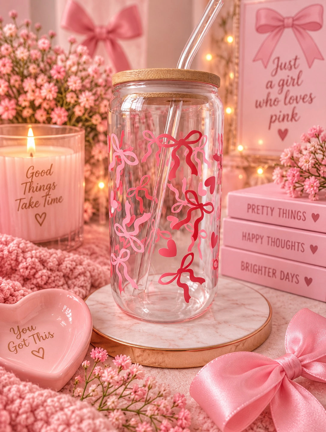 A clear glass mason jar tumbler with a bamboo lid and clear straw, decorated with pink ribbon and heart motifs. It is styled in a romantic pink-themed setting with a pink candle, a pink heart dish, a pink book, and pink flowers.