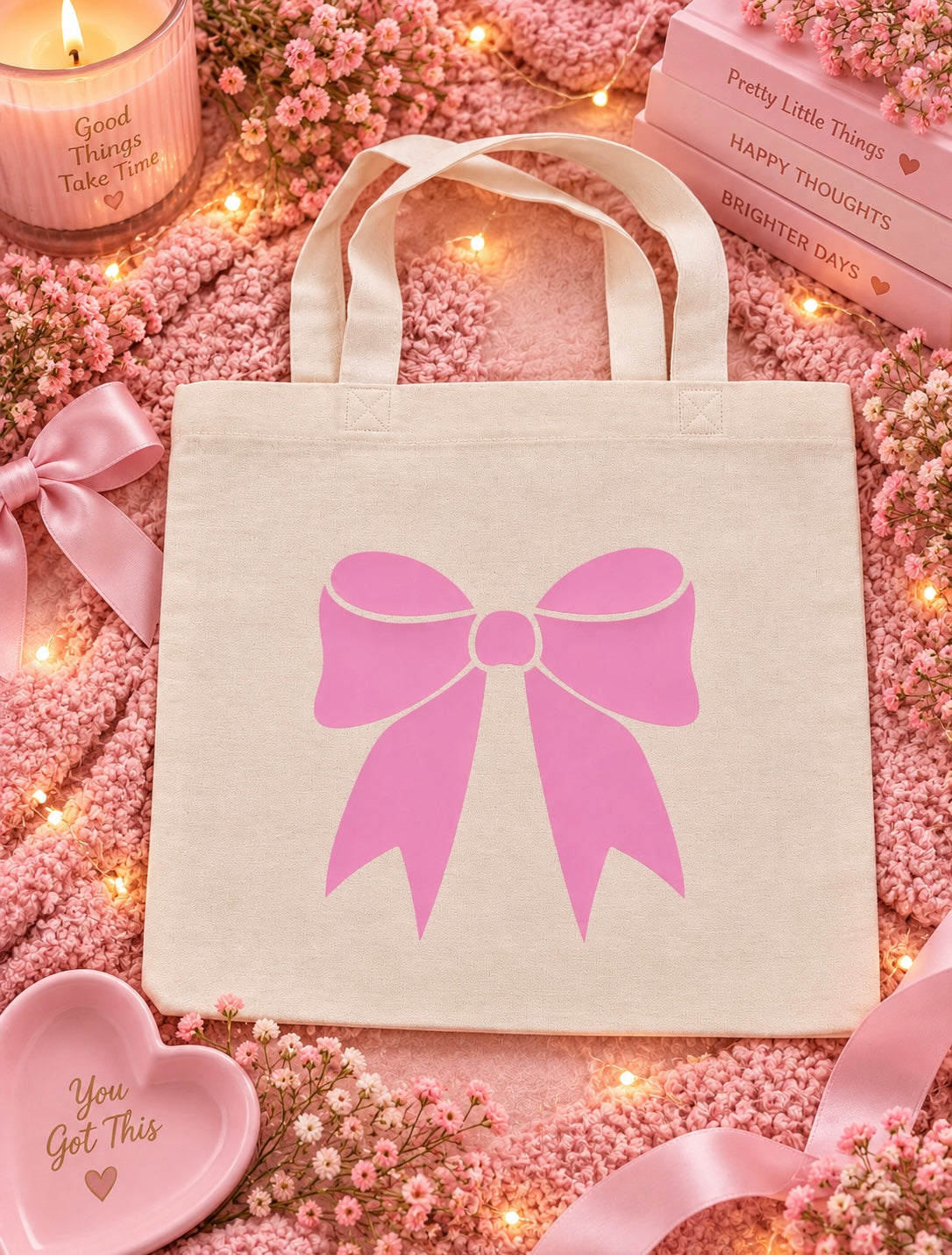 A cream canvas tote bag with a large pink bow graphic on the front, styled in a cozy pink flat-lay with baby's breath flowers, pink ribbons, a candle, and a heart-shaped dish.