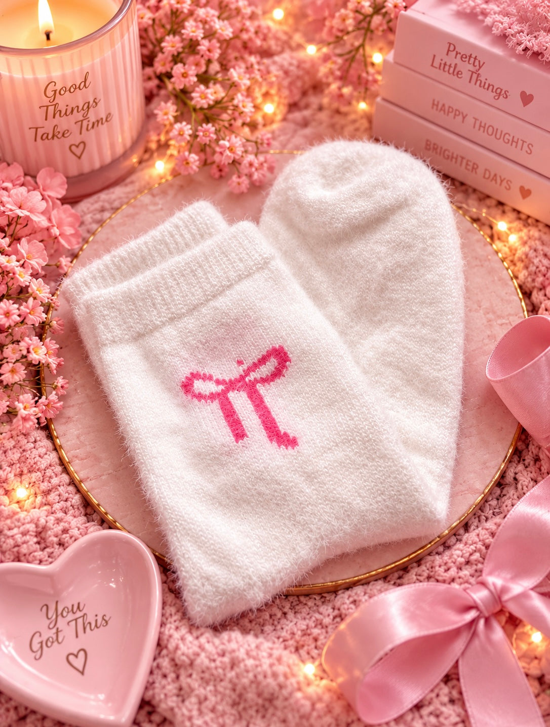 A pair of white knit socks with a pink bow motif on the front, styled in a cozy pink-themed flat lay with flowers, a candle, books, and a pink heart-shaped dish.