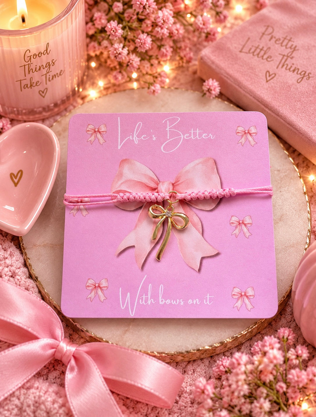 A pink braided bracelet with a bow charm is displayed on a pink presentation card that reads 'Life's Better With bows on it.' The card is styled in a cozy, warm pink setting with dried flowers, a lit candle, and a pink book.