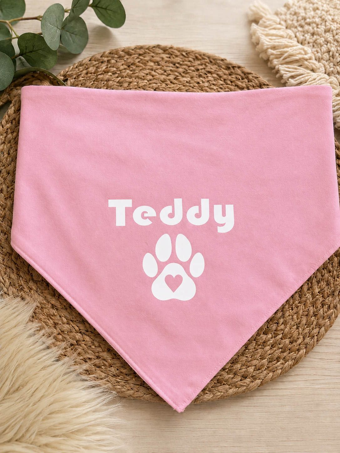 A pink pet bandana with the name 'Teddy' and a paw print graphic printed in white, styled on a woven placemat with decorative greenery and faux fur.