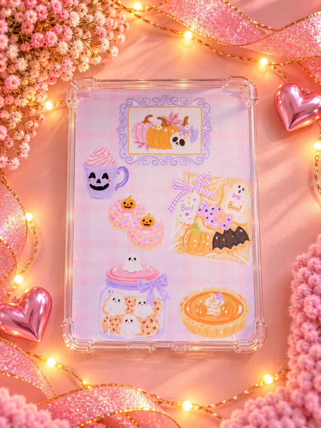 A clear protective Kindle case decorated with cute Halloween-themed insert, including pumpkins, ghosts, cupcakes, and a jar of white ghosts, displayed on a pink background with decorative lights and ribbons.