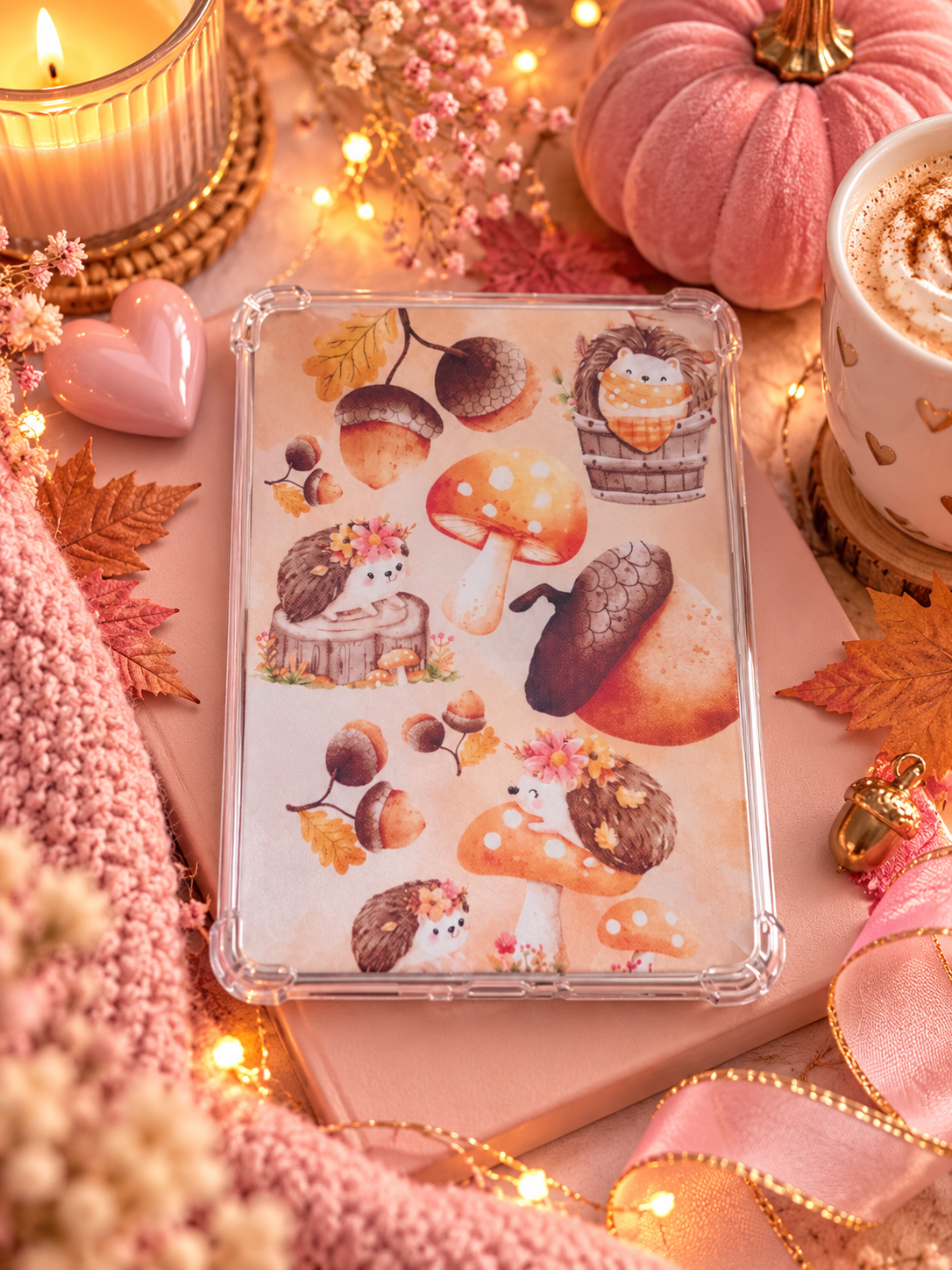 A clear protective kindle case decorated with cute watercolor-style woodland illustrations, including mushrooms, acorns, hedgehogs, and a birdhouse. The case is styled in a cozy autumn-themed setting with a pink knit blanket, fairy lights, a candle, a pink pumpkin, and a cup of hot cocoa.
