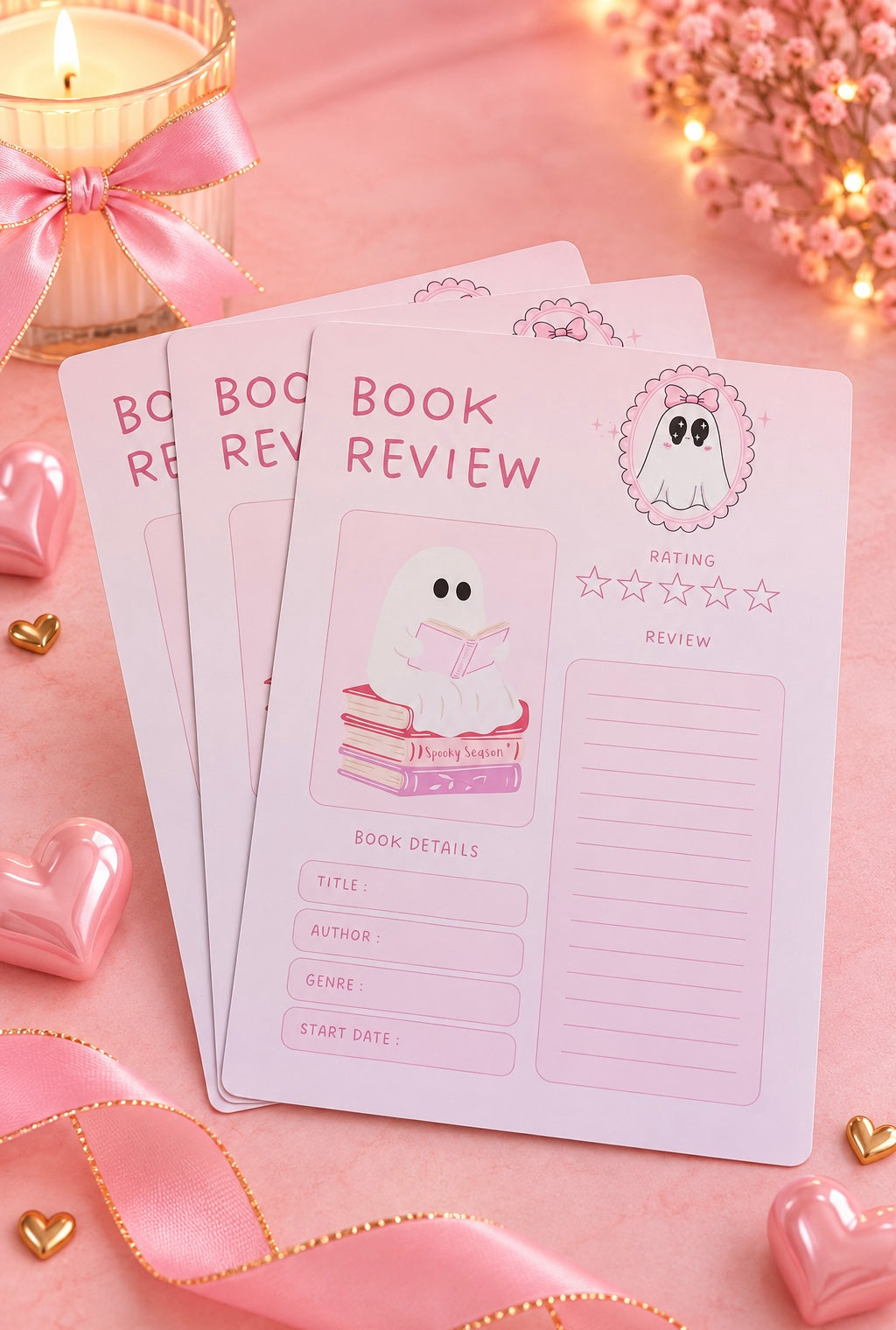 Three pink book review cards with cute ghost illustrations are arranged on a pink surface with decorative hearts, ribbon, and a candle.