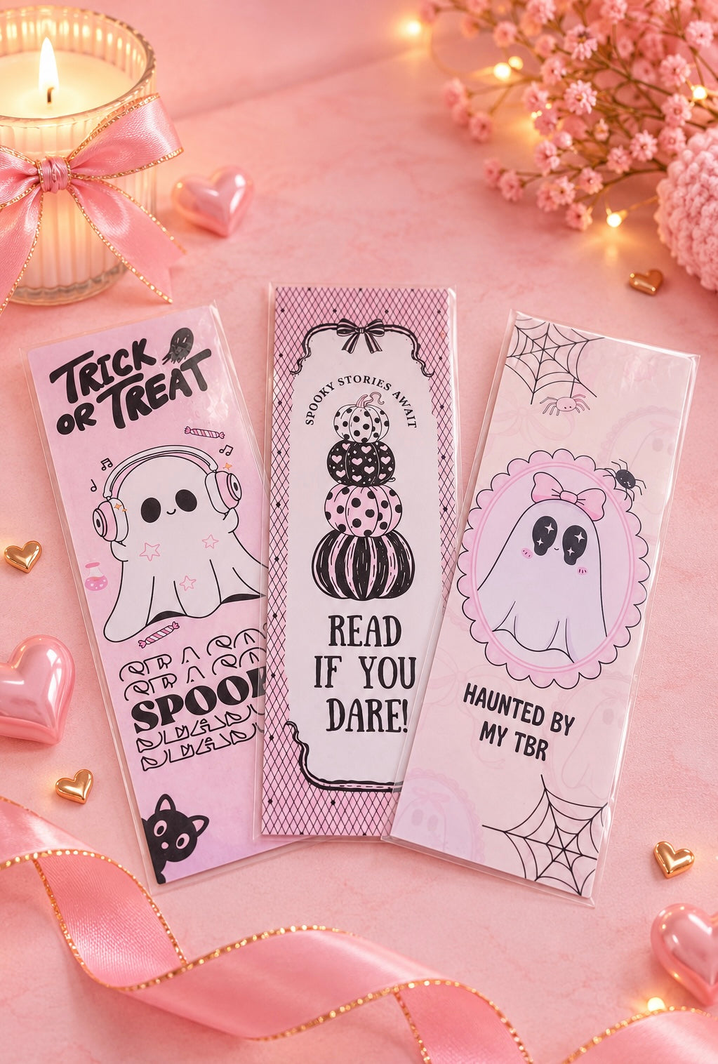 Three pink Halloween-themed bookmarks with cute ghost and pumpkin illustrations, arranged on a pink background with a lit candle, pink ribbon, and small flowers.