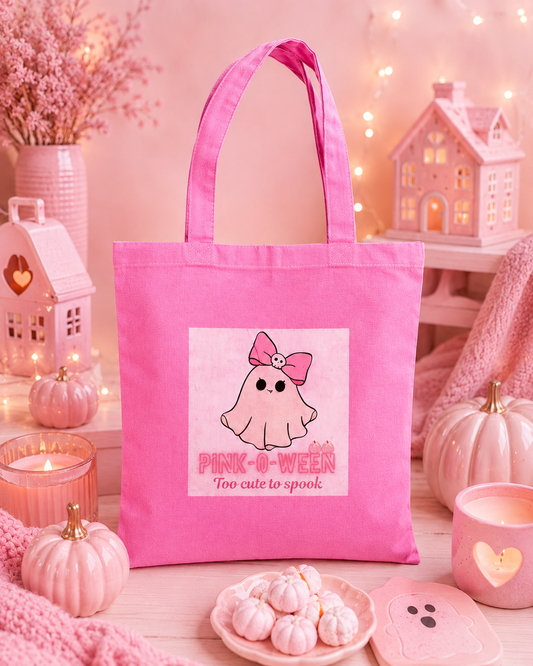 A pink canvas tote bag with a cute ghost graphic and the text 'PINK-O-WEEEN Too cute to spook' displayed in a cozy, fall-themed setting with pumpkins, candles, and a miniature house.