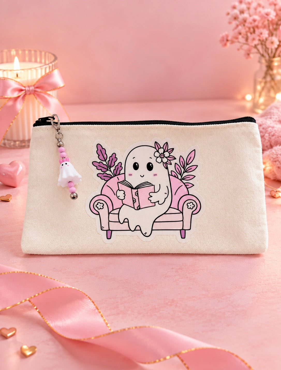A cream-colored canvas zippered pouch featuring a cute cartoon ghost character sitting on a pink sofa and reading a book. The pouch is decorated with a matching charm and is set against a soft pink background with decorative ribbons, a candle, and flowers.