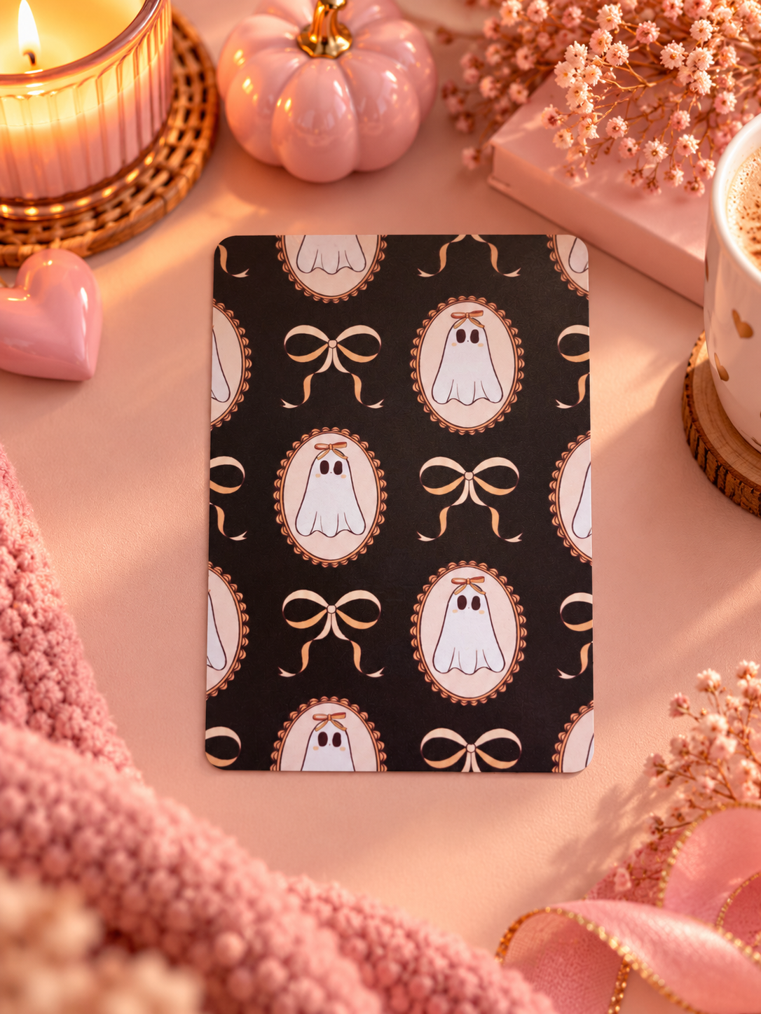 A rectangular kindle insert with a black background and a repeating pattern of white ghosts inside gold-trimmed oval frames, surrounded by pink seasonal props including a candle, small pumpkin, ribbon, and dried flowers.