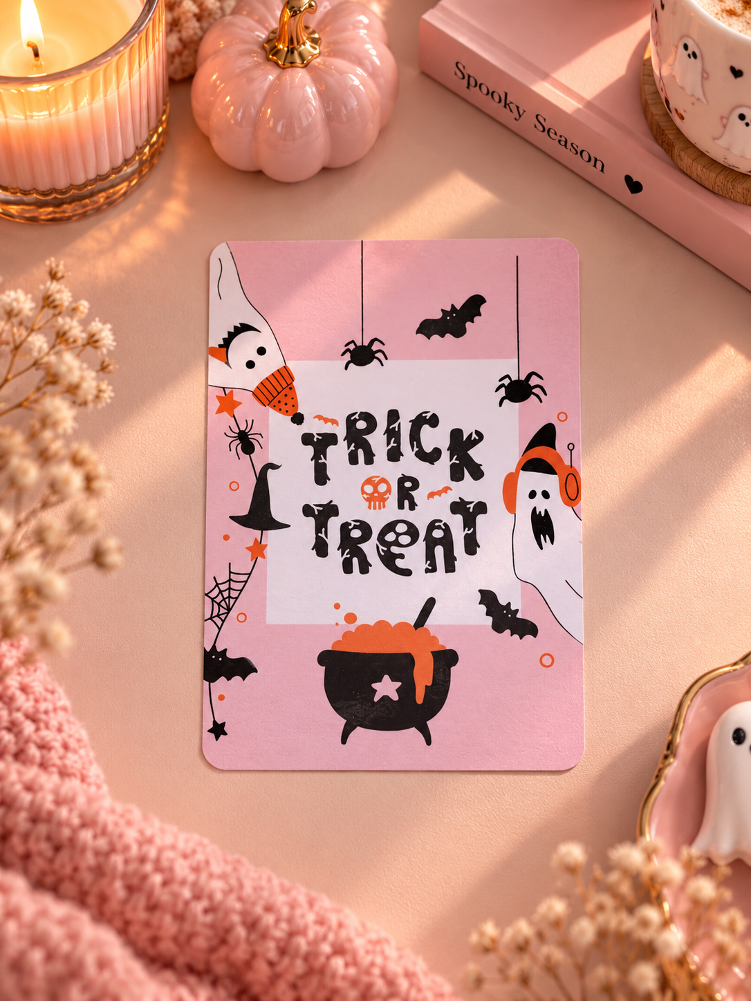 A pink Halloween-themed kindle insert card with 'Trick or Treat' text and spooky illustrations, styled on a pink surface with candles, dried flowers, a small pumpkin, and a book.