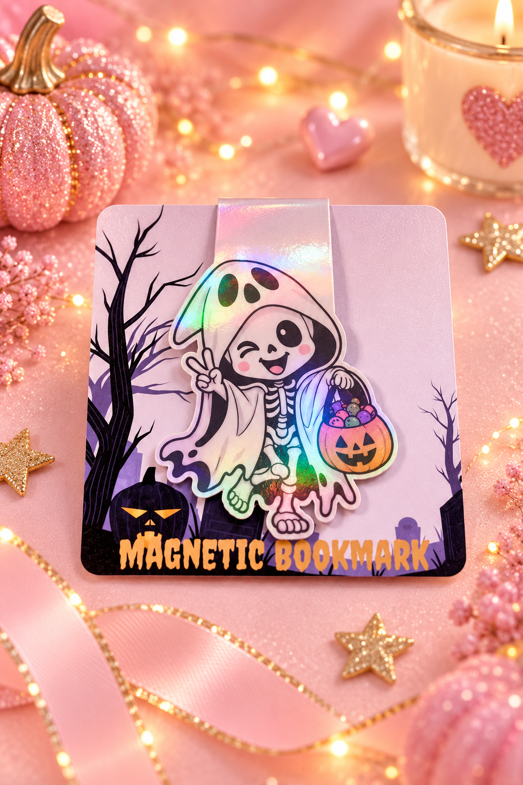 A Halloween-themed magnetic bookmark with a cute skeleton in a ghost cloak holding a pumpkin bucket, displayed on a pink glittery surface with decorative pumpkins, candles, and string lights.