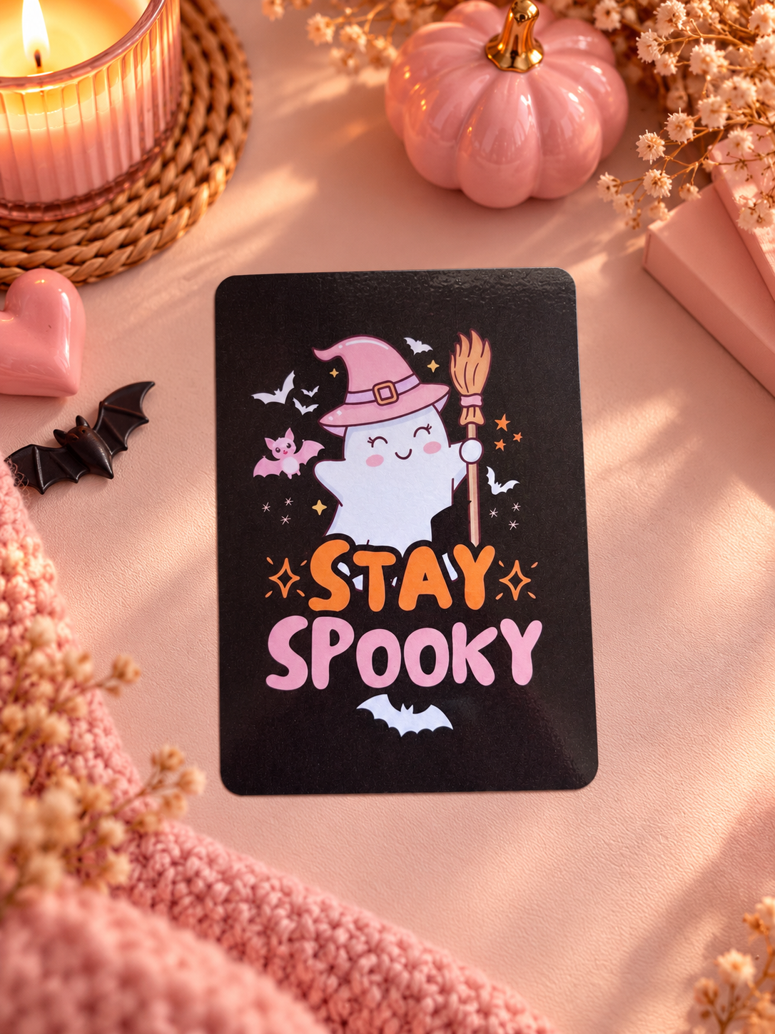 A rectangular black kindle insert with rounded corners featuring a cute cartoon ghost in a pink witch hat holding a broomstick, surrounded by bats and the words “STAY SPOOKY” in orange and pink. The card is styled in a cozy flat-lay with a pink knit blanket, dried flowers, a pink pumpkin, a candle, and a small pink heart on a peach-toned surface.