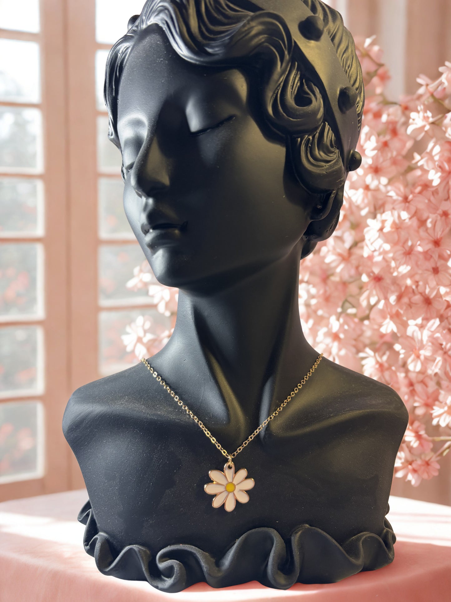 Black bust with a gold necklace featuring a flower pendant against a blurred floral background