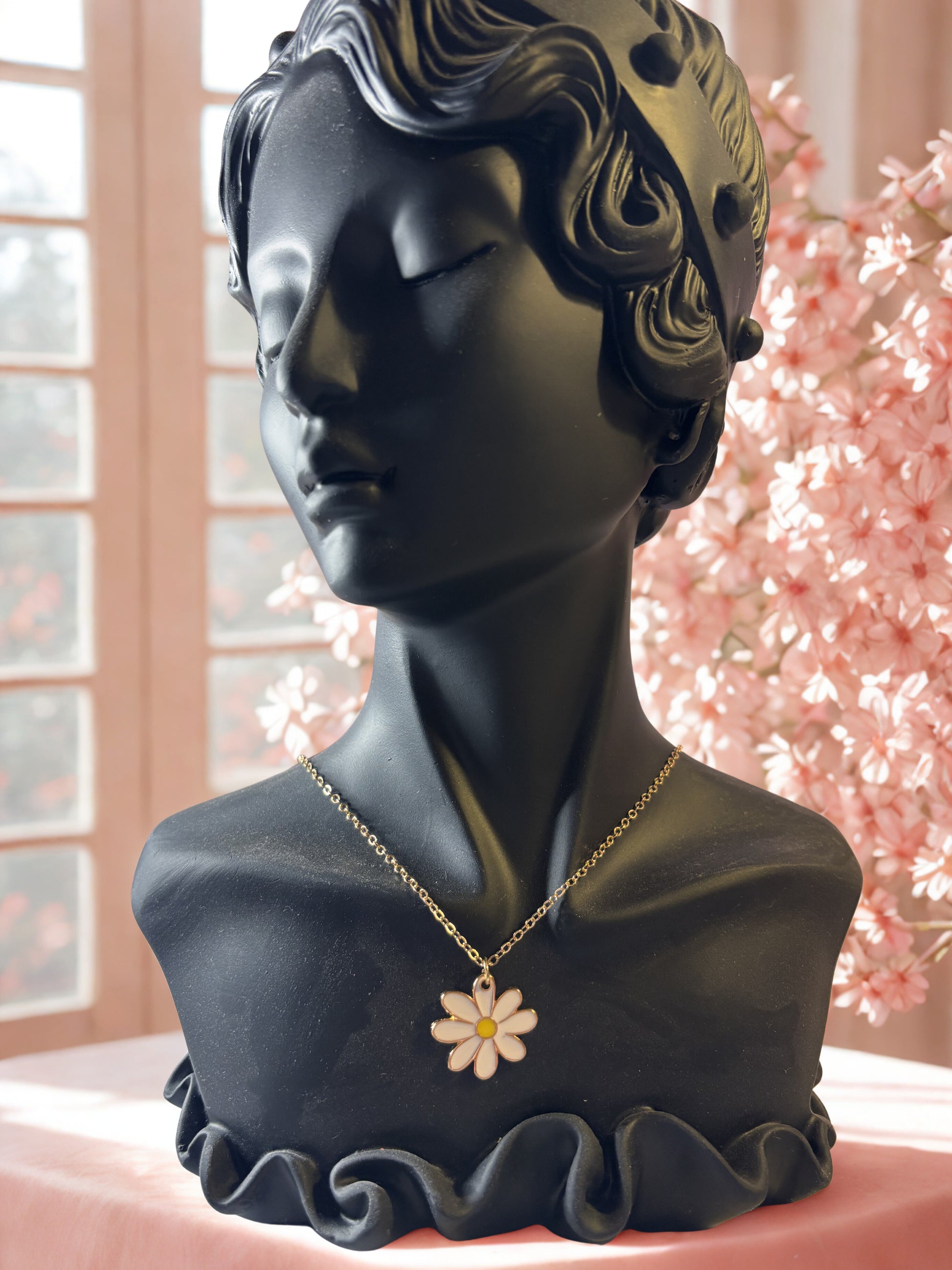 Black bust with a gold necklace featuring a flower pendant against a blurred floral background