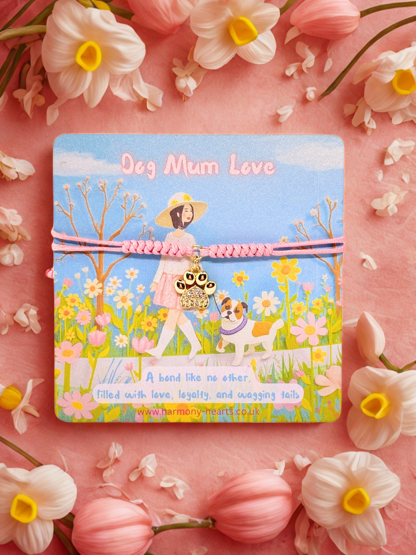 Card with a design of a woman walking a dog in a field, surrounded by flowers on a pink background. With a paw print charm bracelet 