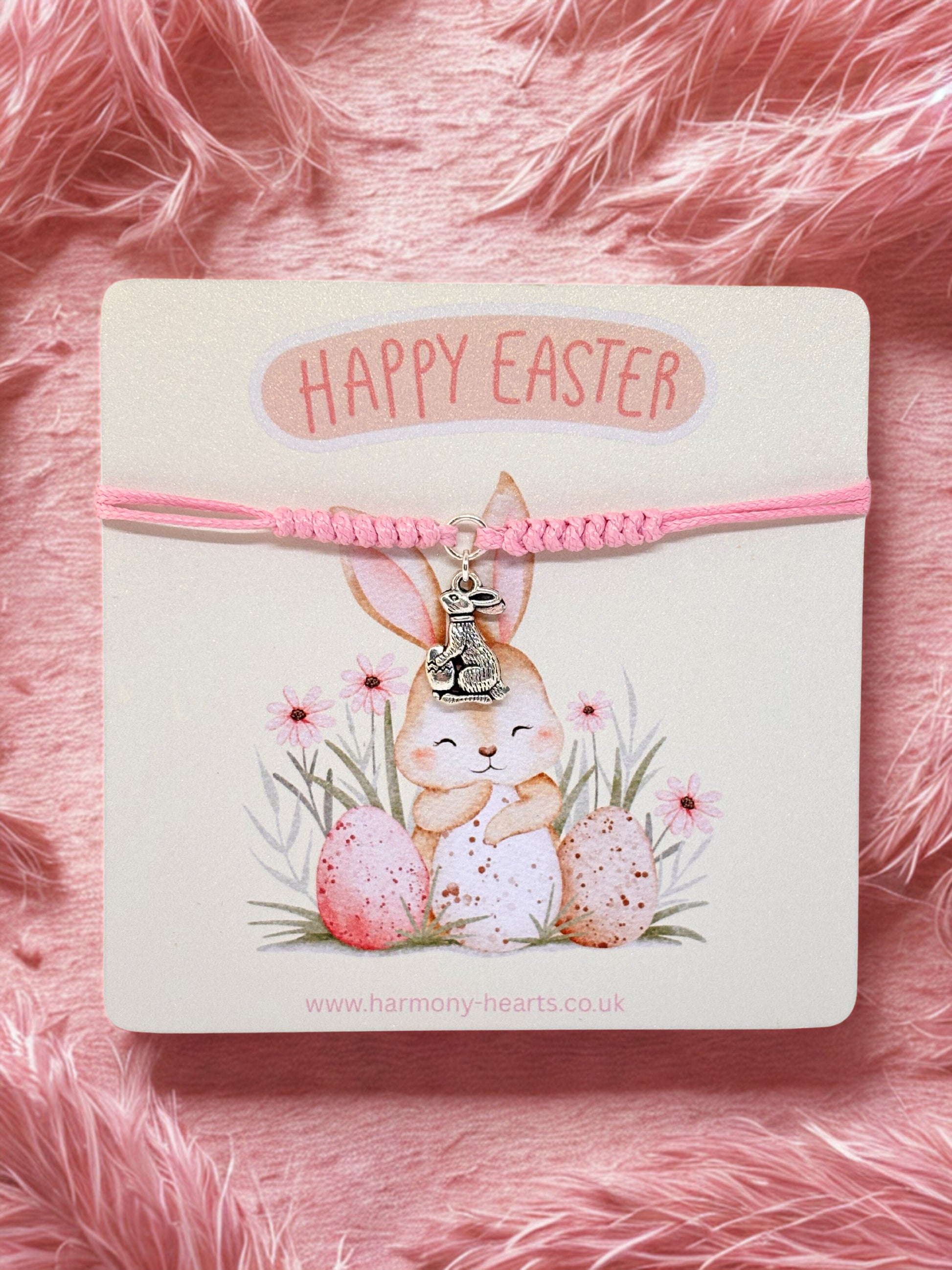Pink bracelet with bunny charm on a card with 'Happy Easter' text, against a pink fluffy background.