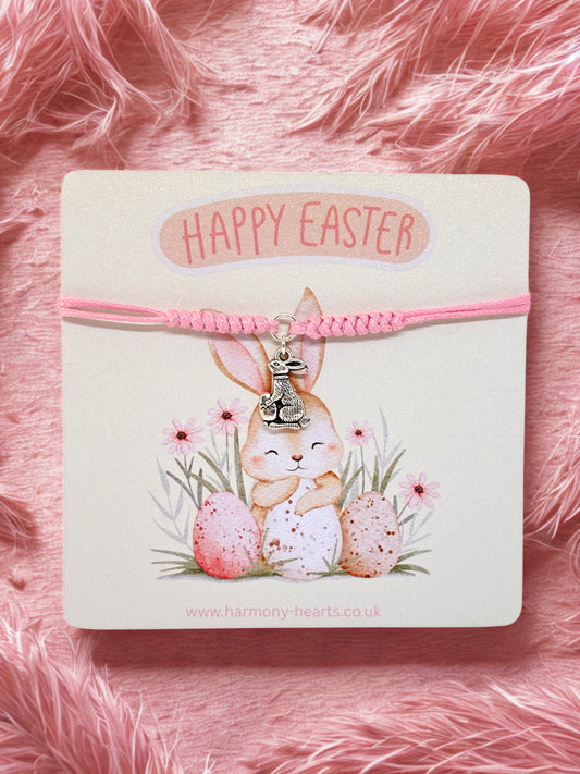 Pink bracelet with bunny charm on a card with 'Happy Easter' text, against a pink fluffy background.