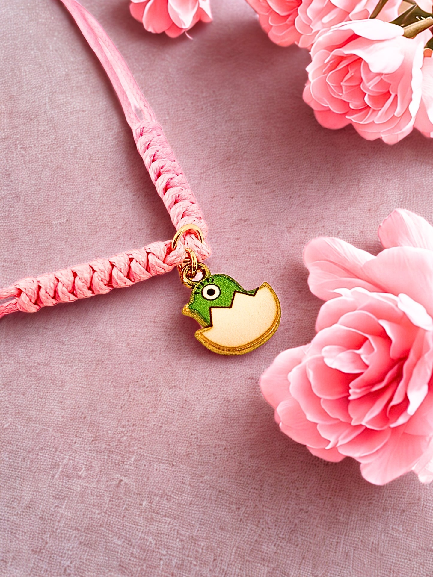 Pink cord bracelet with a green and gold Easter chick charm on a pink fabric background with pink flowers.