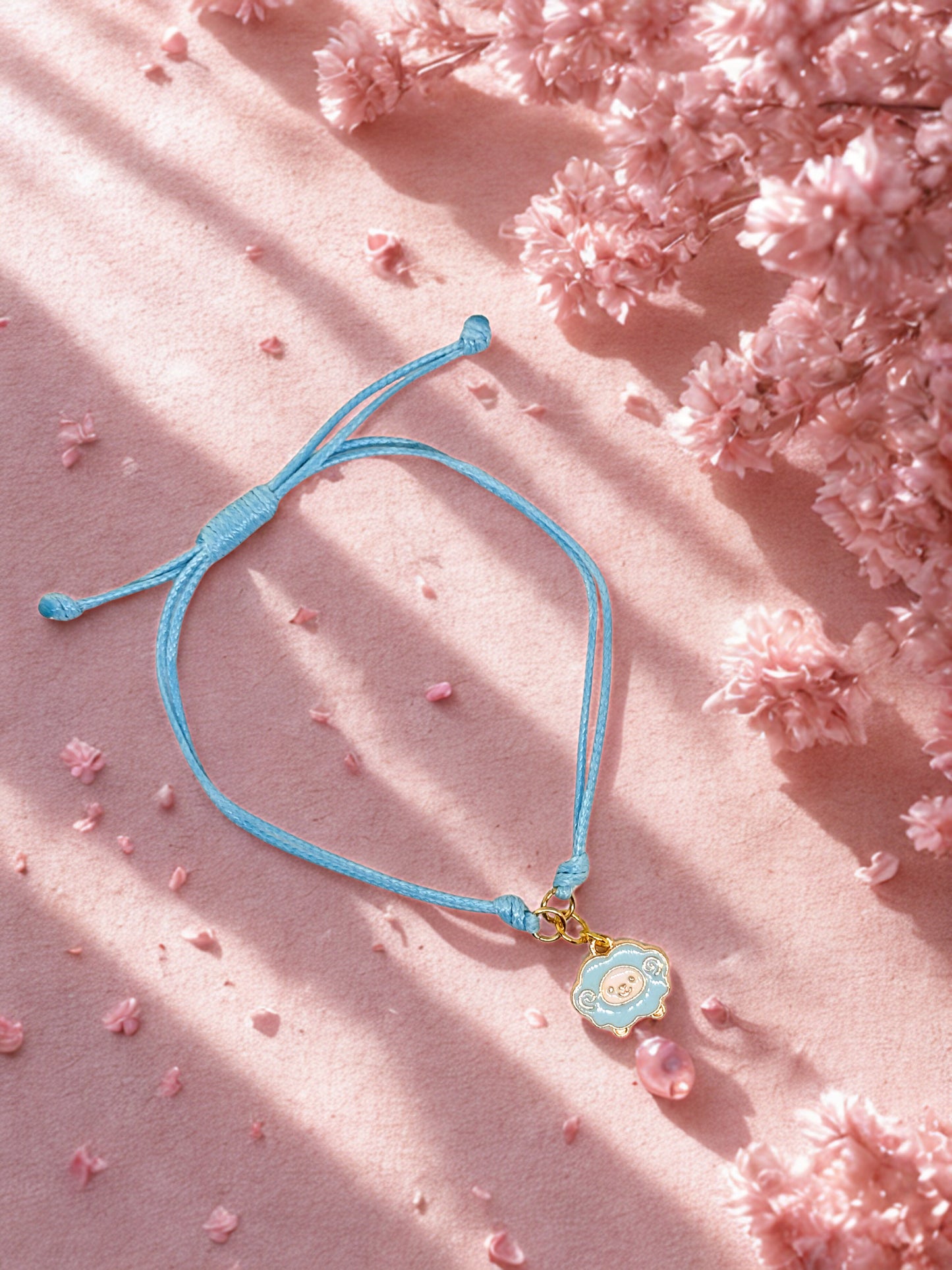Blue cord bracelet with a sheep  charm on a pink surface with cherry blossoms
