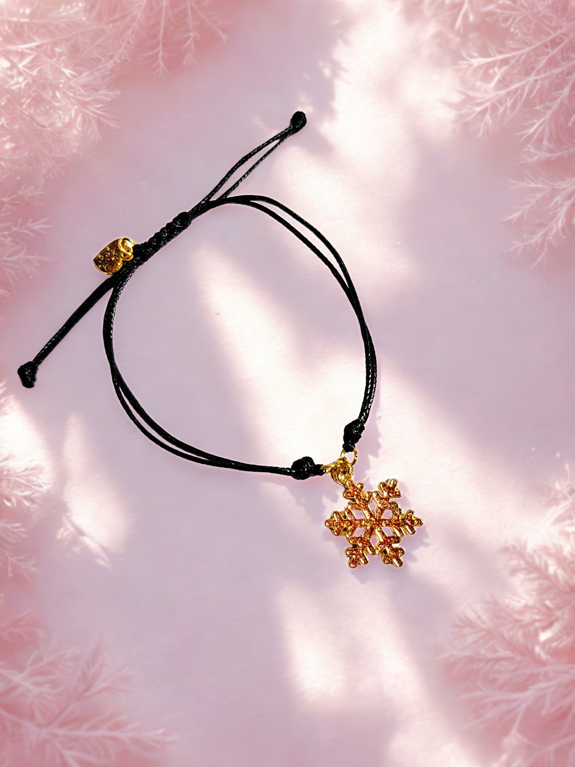 Black cord bracelet with gold snowflake charm on a pink textured background