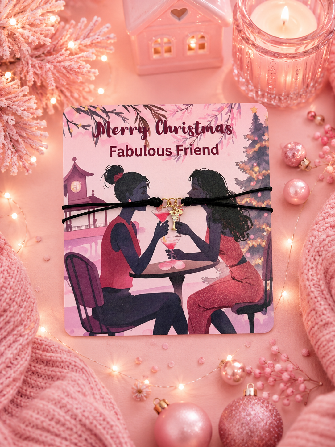 A square gift card featuring a Christmas-themed illustration of two women at a table with the text 'Merry Christmas Fabulous Friend', wrapped with a black cord bracelet and a small gold cocktail charm, styled on a pink holiday background with lights, ornaments, and knit fabric.