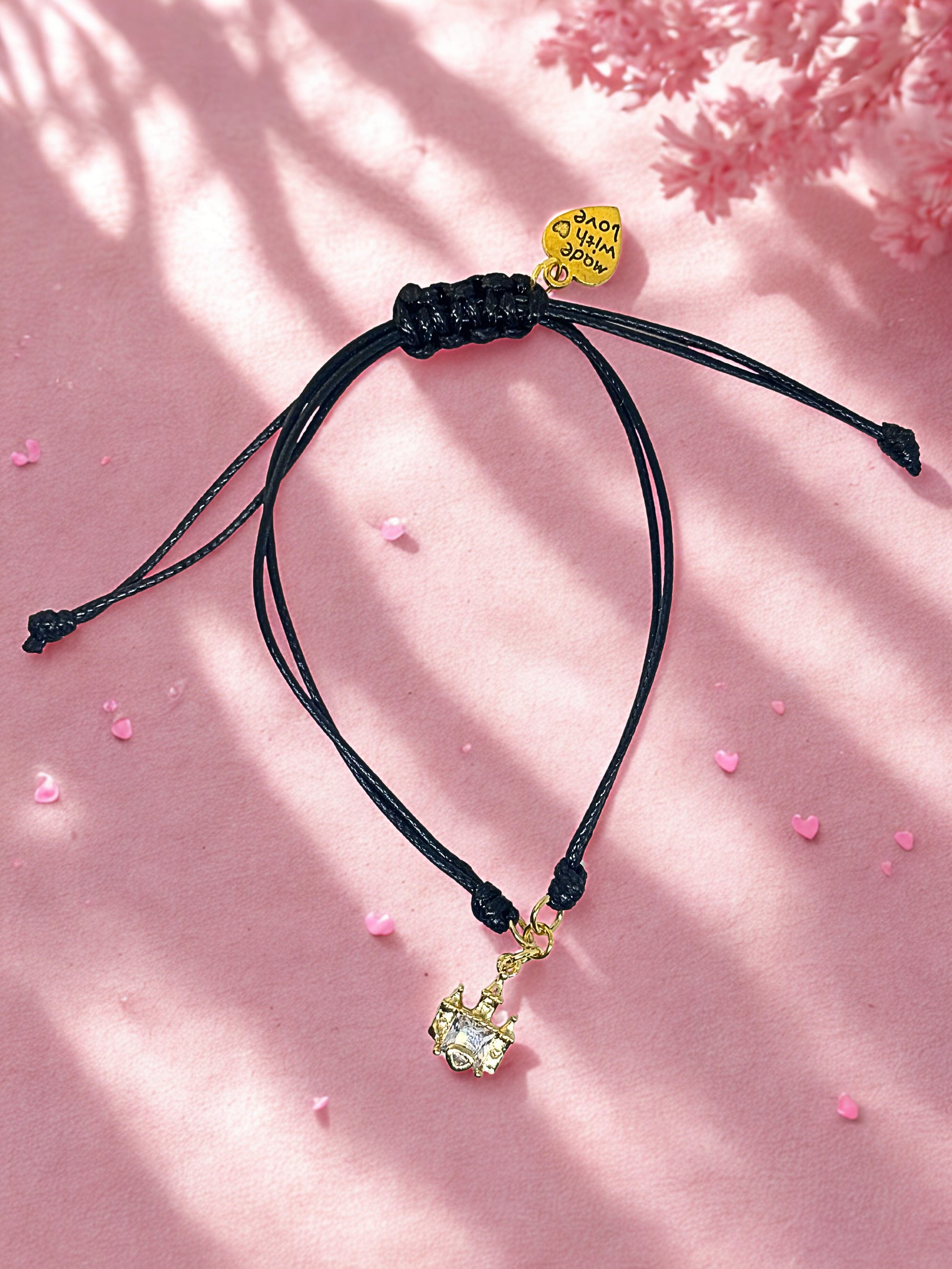 Black cord bracelet with a gold castle charm on a pink background