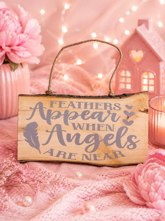 A rustic wooden hanging sign with a twine loop and the phrase 'FEATHERS APPEAR WHEN ANGELS ARE NEAR' in gray lettering, styled on pink fabric with flowers, pearls, fairy lights, and a pink house decoration in the background.