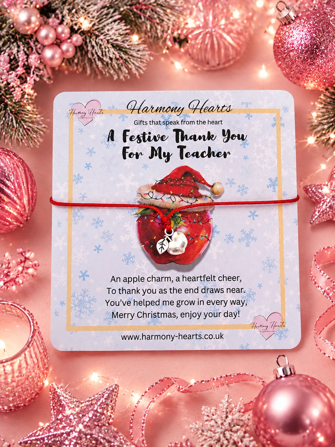 A square card from Harmony Hearts featuring a festive thank you message for a teacher, wrapped with a red string apple charm bracelet. The card is styled in a Christmas flat-lay with pink ornaments, lights, and evergreen branches around it.