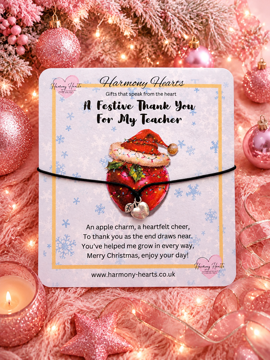 A square gift card from Harmony Hearts wrapped with a black cord apple charm bracelet placed on a festive Christmas background with pink ornaments, lights, and a poem about a teacher. The card features an illustration of a heart-shaped apple wearing a Santa hat.