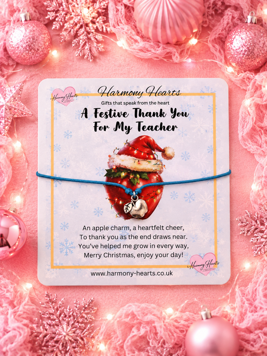 A festive thank you Teacher card from Harmony Hearts wrapped with a blue cord apple charm bracelet, surrounded by pink Christmas ornaments and lights.