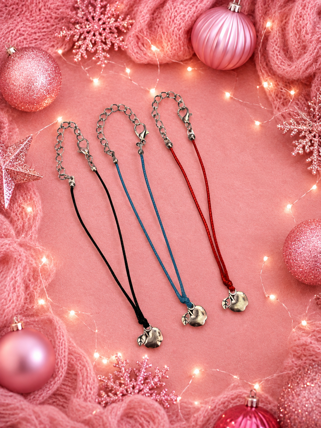 Three charm bracelets with silver-toned apple charms are laid out on a pink background decorated with pink knit fabric, ornaments, snowflake cutouts, and warm string lights. The necklaces have black, blue, and red cords.